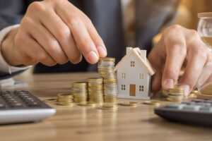 Spekulationssteuer vermeiden beim Immobilienverkauf – Hände stapeln Münzen neben Hausmodell und Sanduhr als Symbol für 10-Jahres-Haltefrist